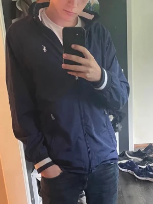 Marinblå vindjacka från Polo Ralph Lauren - Snygg marinblå vindjacka från Polo Ralph Lauren med vit logga på bröstet, dragkedja framtill och huva. Jackan har vita detaljer vid ärmsluten och två fickor med dragkedja. Perfekt för blåsiga dagar och enkel att matcha. Storleken är Xl ungdom motsvarar S