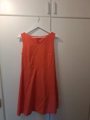 Korall eller orange ärmlös klänning - Snygg orange eller korallfärgas klänning i bomull med polyesterfoder i samma färg. Klänningen är ärmlös, har rund halsringning och en lätt utställd kjol. Dragkedja i ryggen för enkel påklädning. Perfekt för varma dagar och somriga tillfällen.