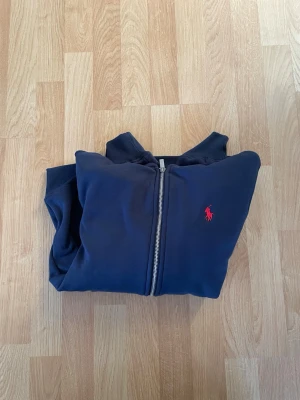 Marinblå hoodie från Polo Ralph Lauren - Snygg marinblå hoodie från Polo Ralph Lauren med dragkedja framtill och vit dragsko i huvan. Klassisk röd logga broderad på bröstet. Tillverkad i mjuk bomullsblandning och har ribbade muddar vid ärmar och nederkant. Perfekt för en avslappnad och stilren look.