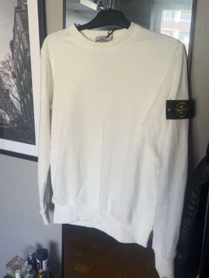 Vit sweatshirt från Stone Island - Vit sweatshirt från Stone Island med klassisk rund hals och ribbade muddar. Tröjan har den ikoniska svarta logotypen med kompass på vänster ärm. Perfekt för en clean och stilren look.