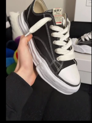 Svarta MMY sneakers med vit sula - Snygga svarta sneakers från Maison Mihara Yasuhiro med vit, chunky sula och kontrasterande vita sömmar. Skorna har breda vita skosnören och en unik, vågig sula som sticker ut. Ovandelen är i canvas och tån är vit. Perfekt för dig som gillar streetwear och statement sneakers.