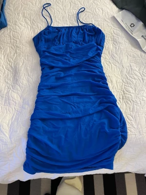 Blå kort klänning med tunna axelband - Snygg blå kort klänning med tunna axelband och rynkade detaljer längs hela klänningen. Figurnära passform och stretchigt material som ger en smickrande siluett. Perfekt för dig som vill sticka ut med en stark färg.