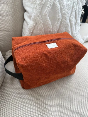 Rostfärgad necessär från Emelo - Necessär i rostfärgat lent möbeltyg från Emelo. Rektangulär form med dragkedja upptill och svart bärhandtag på sidan. Märke på framsidan. Perfekt för smink eller småprylar. Tygmaterial med enkel, stilren design. Beige insida med innerficka. Storlek large. Mått ca 27x17x12cm