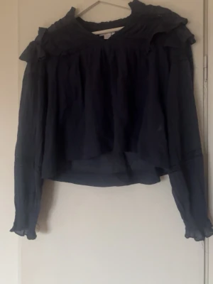 Mörkblå volangblus med lång ärm - Snygg mörkblå blus i tunn, luftig kvalitet med volangdetaljer vid axlarna och rynkade muddar vid ärmsluten. Blusen har en lös passform och transparent tyg som ger en trendig look. Perfekt att styla med jeans eller kjol.