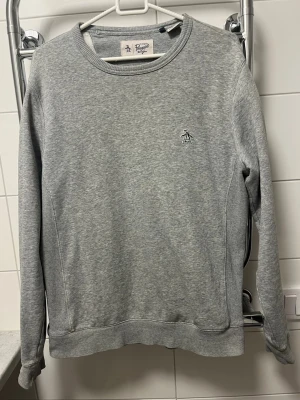 Original pinguin - Klassisk grå sweatshirt från Original Penguin med rund halsringning och diskret broderad pingvinlogga på bröstet. Tröjan har långa ärmar, ribbade muddar och är tillverkad i mjukt bomullsmaterial. Enkel och stilren design som funkar till det mesta.