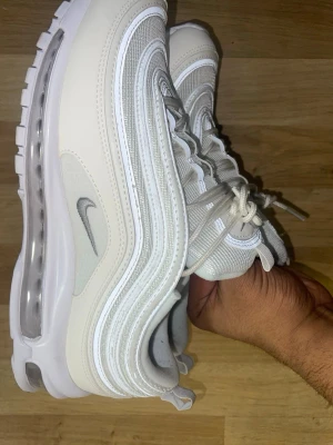 Nike Air Max 97  - Nike Air Max 97 sneakers i vitt och ljusgrått med ikoniska vågiga paneler och reflekterande detaljer. Ovandelen är i mesh och syntet, med synlig Air Max-sula för extra komfort. Klassisk Swoosh-logga på sidan och snörning framtill.