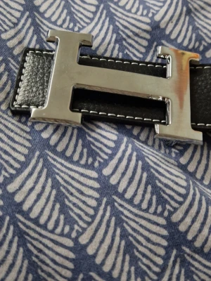 Svart skärp med silver H-spänne - Snyggt svart skärp från H med ett stort H-format spänne i silverfärg. Skärpet är gjort i slätt skinn med vita sömmar längs kanterna. Perfekt accessoar för att lyfta vilken outfit som helst.