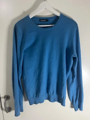 Blå stickad tröja från J.Lindeberg - Snygg blå stickad tröja från J.Lindeberg med rund halsringning och diskret logga broderad på bröstet. Tröjan har långa ärmar och ribbade muddar vid ärmslut och nederkant. Perfekt till lager på lager och enkel att matcha.