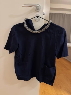 Mörkblå topp med pärldetaljer från ZARA - Snygg mörkblå topp från ZARA med korta ärmar och två fickor framtill. Kragen är dekorerad med flätade pärldetaljer i silver och svart som ger en unik touch. Perfekt för dig som vill sticka ut lite extra.
