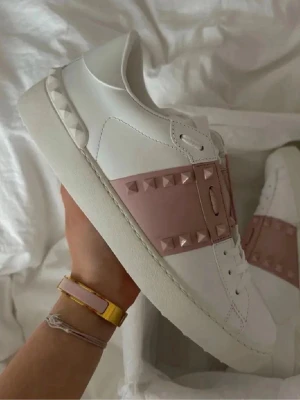Valentino Garavani sneakers med nitar - Snygga vita sneakers från Valentino Garavani med bred rosa rem över foten dekorerad med ikoniska pyramidformade nitar. Skorna har en klassisk låg siluett, vita skosnören och detaljer i läder. Perfekta för dig som vill ha en unik touch på din outfit.