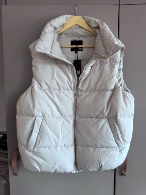 Vit dunväst från Lager 157 - Säljer en ljusgrå dunväst från Lager 157 med hög krage och dragkedja framtill. Västen har två snedställda fickor och quiltad design som ger en puffig look. Perfekt för lager på lager och riktigt snygg till streetwear-stilen.