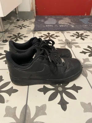 Nike Air Force 1 Low helsvarta sneakers - Säljer ett par klassiska Nike Air Force 1 Low sneakers i helsvart läder med svart sula och svarta skosnören. Ikonisk design med Nike-logga på sidan och bak. Perfekta för dig som vill ha en clean och tidlös look.
