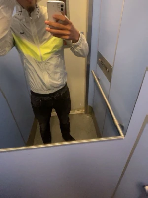 Lacoste vindjacka med gul detalj - Säljer en snygg vindjacka från Lacoste i ljusbeige och grått med en skarp gul V-formad detalj över bröstet. Jackan har dragkedja, huva med snörning och Lacoste-loggan på vänster sida. Materialet är lätt och perfekt för att skydda mot vind. Perfekt för sommar och vår, den är använd 1-2 gånger. 