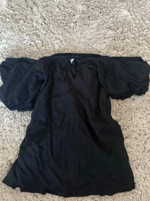 Svart offshoulder blus med puffärm - Säljer en svart offshoulder siden blus med breda puffärmar och smockdetaljer vid axlarna. Blusen har en lös passform och ett litet knyt framtill vid halsen. Perfekt för dig som gillar trendiga och bekväma toppar.