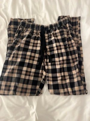 Rutiga pyjamasbyxor från Ben Barton - Mysiga pyjamasbyxor från Ben Barton i beige, svart och vitt rutigt mönster. Byxorna har resår i midjan med snörning för extra komfort. Perfekta för chillkvällar hemma och tillverkade i mjukt tyg som känns skönt mot huden.