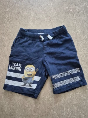 Shorts - Säljer ett par mörkblå shorts med Minion-tryck och texten 'Team Minion' på vänster ben. Shortsen har vita och blå ränder, resårmidja med snörning och är gjorda i mjuk bomull. Perfekta för sommarens alla äventyr!