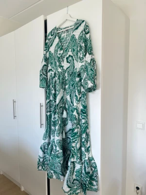 Grön paisleymönstrad långklänning Lindex - Långklänning från Lindex i vitt och grönt med paisleyinspirerat mönster. Klänningen har v-ringning, volangdetaljer nertill och trekvartsärm. Luftig passform och tillverkad i mjuk bomull, perfekt för varma dagar.