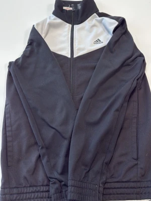 Svart och vit träningsjacka Adidas - Snygg svart träningsjacka från Adidas med vit panel upptill och dragkedja hela vägen. Jackan har hög krage, två sidofickor och Adidas-logga på bröstet. Tillverkad i ett mjukt och stretchigt material som passar perfekt till träning eller sport.