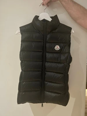 Svart dunväst från Moncler - Snygg svart dunväst från Moncler med hög krage och quiltad design. Västen har dragkedja framtill och Monclers ikoniska logotyp på bröstet. Perfekt för lager-på-lager och ger en clean streetstyle-look. ÄKTA! Nypris - 9000-13000