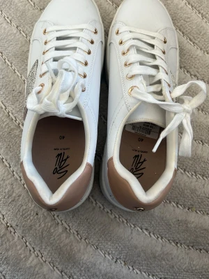 Vita sneakers med guld och strass - Snygga vita sneakers från SO med guldfärgade detaljer och strass på sidan. Skorna har vita snören, tjock sula och beige/guld hälparti. Perfekta för dig som vill ha en trendig och bekväm look. Materialet är konstläder. Sulan är ganska så hög, använda 1 gång 