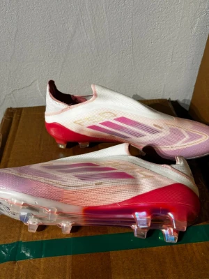 Adidas vita och rosa fotbollsskor - Säljer ett par Adidas fotbollsskor i vitt och rosa med röda detaljer. Skorna har en strömlinjeformad design utan snörning och genomskinliga dobbar för bästa grepp på planen. Tillverkade i syntetmaterial för lätt känsla och snabbhet.