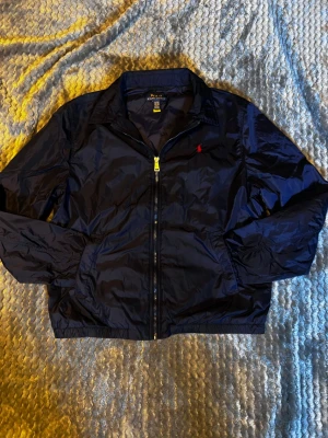 Mörkblå vindjacka från Polo Ralph Lauren - Snygg mörkblå vindjacka från Polo Ralph Lauren med klassisk krage, dragkedja framtill och två sidofickor. Jackan har elastiska muddar vid ärmslut och nederkant samt den ikoniska röda logotypen broderad på bröstet. Tillverkad i lätt och glansigt material.