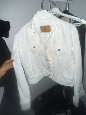 Vit jeansjacka från H&M - Säljer en snygg vit jeansjacka från H&M med klassisk design, två bröstfickor med knappar och silverfärgade detaljer. Jackan har en rak passform och är tillverkad i jeansmaterial. Perfekt att slänga över en t-shirt för en cool look. 