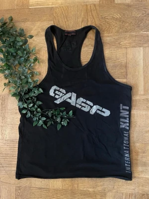 Svart linnetopp från GASP - Säljer en svart linnetopp från GASP med tryck i vitt och grått på framsidan. Modellen är ärmlös med djupa ärmhål och brottarrygg, perfekt för träning. Materialet känns mjukt och stretchigt, 