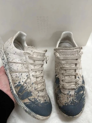Maison margiela - Unika beige sneakers med blå och svarta färgstänk över hela skon. Lågt skaft och klassisk snörning. Tillverkade i canvasmaterial med gummisula. Snyggt sliten look och cool design som sticker ut. Säljer pga att dem är för små skriv om du är intresserad😁
