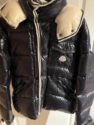 Svart glansig dunjacka från Moncler - Snygg svart glansig dunjacka från Moncler med vita detaljer och avtagbar huva. Jackan har en bröstficka med Moncler-logga, dragkedja framtill och vadderad design som håller dig varm under kalla dagar. Perfekt för dig som vill ha både stil och funktion.