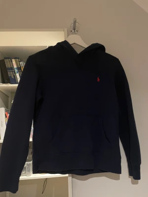 Mörkblå hoodie från Polo Ralph Lauren - Mörkblå hoodie från Polo Ralph Lauren med klassisk röd logga broderad på bröstet. Hoodien har känguruficka framtill, mjukt material och en skön huva. Perfekt för dig som gillar stilrena och tidlösa plagg. Passar 152-158