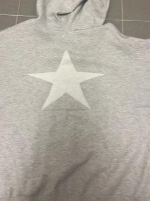 Grå hoodie med vita stjärnor - Snygg grå hoodie med huva och dragsko. På ryggen finns en stor vit stjärna och framtill flera mindre vita stjärnor. Tillverkad i mjukt sweatshirtmaterial som är skönt att ha på sig. Perfekt för en avslappnad och trendig look.