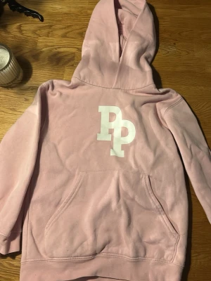 Ljusrosa hoodie med Pidde P tryck - Säljer en ljusrosa hoodie med stor vit Pidde P logga på bröstet. Hoodien har en klassisk känguruficka framtill och en rymlig huva. Storlek: 7-8 år, 128