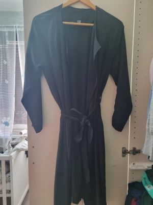 Svart lång kappa från 157 - Stilren svart lång kappa från 157 med knytskärp i midjan och öppen front. Kappan har långa ärmar och en enkel, rak passform som ger en clean look. Perfekt för dig som gillar minimalistisk stil och vill ha en lättare kappa till vardags.
