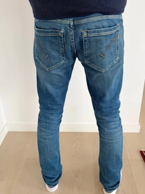 Blå slim fit jeans från dondup - Säljer ett par blå slim fit jeans med snygga slitningar och dragkedjedetaljer på bakfickorna. Har ett litet hål vid ljumsken. Jeansen har klassisk femficksdesign och normal midja. Perfekta för dig som gillar en smalare passform och en modern look.