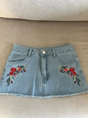 Blå jeanskjol med broderade rosor - Snygg ljusblå jeanskjol med broderade röda rosor och gröna blad framtill. Kjolen har rå kant nertill, klassiska fickor fram och bak samt bälteshällor. Skriv för mått