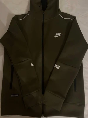 Olivgrön Nike hoodie med dragkedja - Snygg olivgrön hoodie från Nike med hel dragkedja framtill och vit logga på bröstet och ärmen. Hoodien har huva, svarta dragkedjor på fickorna och vita detaljer vid axlarna. Tillverkad i ett mjukt och stretchigt material som känns sportigt och modernt.    Storlek 170