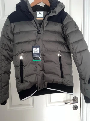 Grå dunjacka med svarta detaljer - Snygg grå dunjacka från Mountain Works med svarta axelpartier och detaljer. Jackan har huva, dragkedja framtill, två fickor med dragkedja och ribbade muddar. Perfekt för kalla vinterdagar och riktigt skön att bära. Storlek 158! Skick 10/10 helt ny! Skriv till mig vid intresse eller frågor!