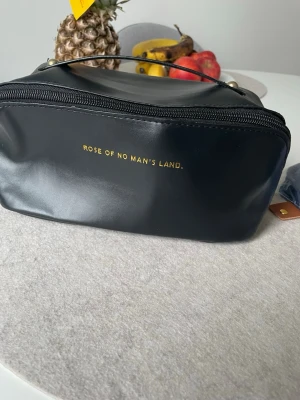 Svart necessär Rose of No Man's Land - Snygg svart necessär i glansigt material med guldfärgad text på framsidan. Väskan har dragkedja, ett praktiskt handtag och ljus insida med flera fack för organisering. Perfekt för smink eller småprylar när du är på språng.