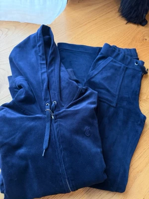 Mörkblå velourset med hoodie - Mysigt mörkblått loungewear-set i mjuk velour med matchande hoodie och byxor. Hoodien har dragsko och dragkedja framtill samt diskret broderad logga på bröstet. Byxorna har resår i midjan och fickor. Perfekt för chill dagar hemma. Byxorna är XSS och koftan Xs. 