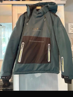 Grön och svart skiddjacka Evil Wolf - Snygg skidjacka från Evil Wolf i mörkgrönt och svart med gula detaljer på dragkedjorna. Jackan har stor ficka framtill, huva med dragsko och muddar vid ärmslut. Perfekt för skidåkning i kallare väder så den är tjock och riktigt varm. Aldrig använd då det blev storlek. 