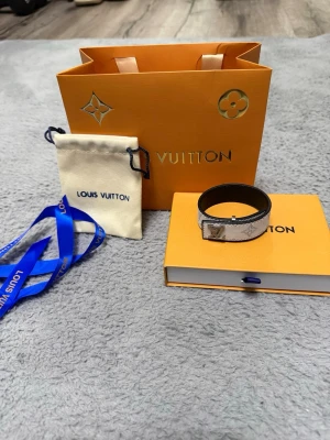 Louis Vuitton armband monogram - Snyggt armband från Louis Vuitton med klassiskt monogrammönster i vitt och beige. Armbandet har en guldfärgad LV-logga på framsidan och är tillverkat i läder med metall-detaljer. Perfekt för dig som gillar exklusiva accessoarer och ikonisk design.