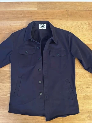 Mörkblå overshirt  - Snygg mörkblå overshirt från Stilgruvan med klassisk krage, knäppning framtill och två bröstfickor med lock och knapp. Tillverkad i ett slitstarkt bomullstyg och har två sidofickor. Perfekt lager på lager-plagg för en clean och stilren look.