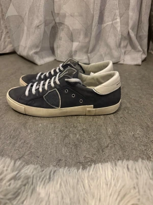 Mörkblåa Philippe Models - Snygga mörkblåa äkta Philippe Models med vita detaljer och vita skosnören. Skorna har en låg profil, klassisk siluett och är tillverkade i mocka och läder. Yttersulan är vit och ger en fräsch kontrast till den mörka ovandelen. Perfekta för en stilren och avslappnad look.