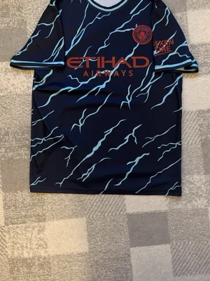 Manchester City matchtröja blå - Säljer en blå Manchester City matchtröja med ljusblå blixtmönster och röda loggor. Tröjan har rund hals och är tillverkad i lätt, ventilerande polyester. Perfekt för fotboll eller träning, med Etihad Airways och Nexen Tire tryck på framsi