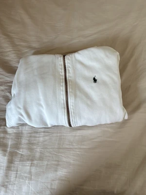 Vit hoodie från Ralph Lauren - Säljer en vit hoodie från Ralph Lauren med dragkedja framtill och klassisk logga på bröstet. Hoodien har huva, fickor på sidorna och ribbade muddar vid ärmslut och nederkant. Perfekt för en clean och stilren look.