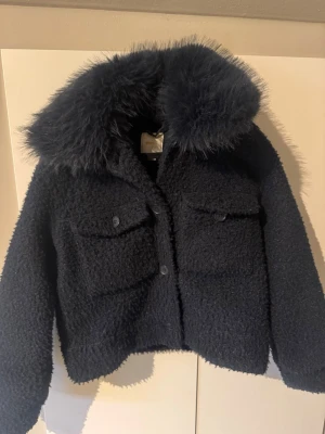 Svart teddyjacka  - Mysig svart teddyjacka med stor fluffig fuskpäls på kragen. Jackan har två stora bröstfickor med knappar och stängs framtill med knappar. Kortare modell som ger en trendig look och passar perfekt till kalla dagar.