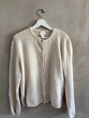 Beige kofta med rund hals från H&M - Mysig beige kofta från H&M med rund halsringning och knäppning framtill. Koftan har långa ärmar, ribbade muddar och är tillverkad i ett mjukt, fluffigt material som känns skönt mot huden. Perfekt för lager på lager och enkel att matcha.