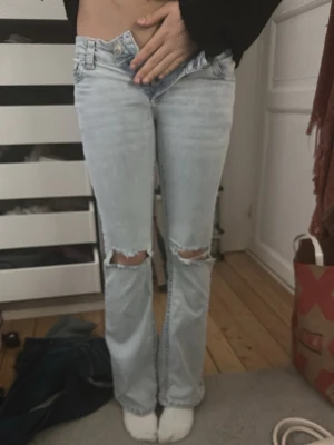 Ljusblå bootcut jeans med slitningar - Säljer ett par ljusblå bootcut jeans med slitna hål på knäna och broderade detaljer i rosa  på bakfickorna. Jeansen har låg midja och klassisk femficksdesign🫰 säljer på grund av att de är för korta och för små❤️pris kan diskuteras❤️
