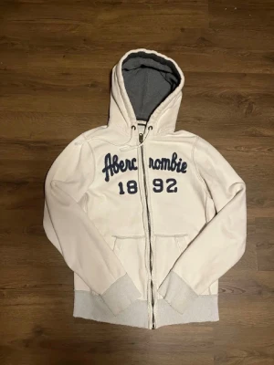Vit Abercrombie & Fitch hoodie fint skick - Snygg vit hoodie från Abercrombie & Fitch med dragkedja och mörkblå text. Mjuk insida, ribbade muddar och fickor framtill. Är XL fast passar som en L. Är själv 190 och den passar super👌 OBS! Alla slitage på bilden är fabriks gjorda!👏
